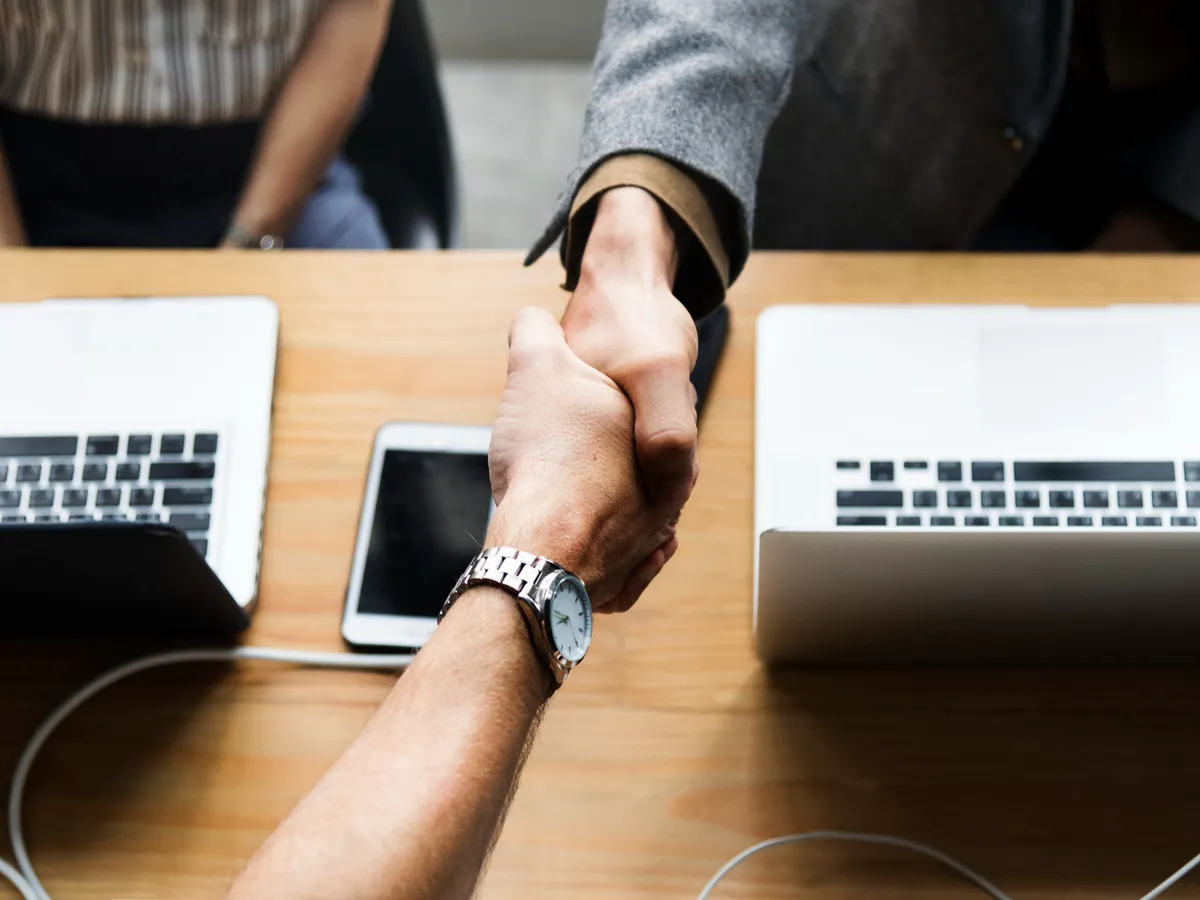 A handshake over a conference table, with two laptops and a phone in the background